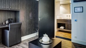 A modern hotel room with a dark wood panel wall, a black desk with a kettle, a chair, and a decorative white sphere on a black tray.