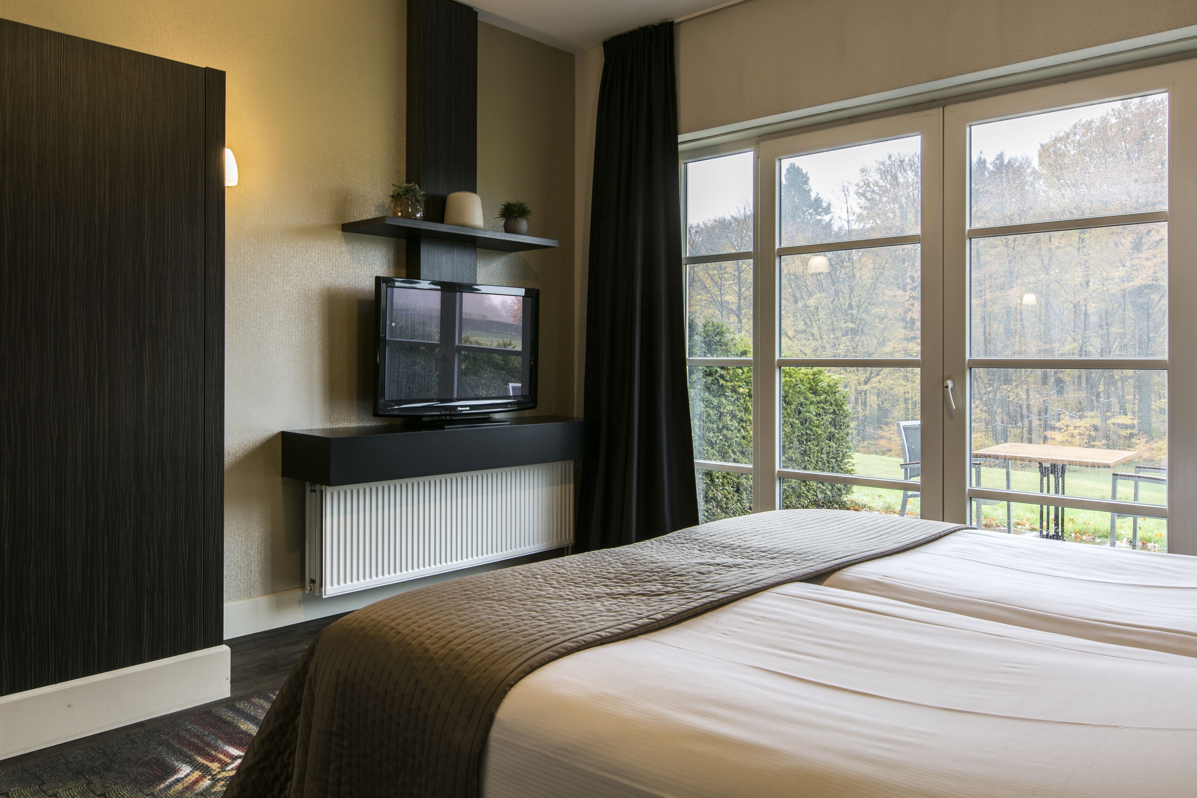 Un dormitorio con una cama grande, un televisor sobre un soporte negro, una ventana con vistas a árboles y un radiador debajo del televisor.