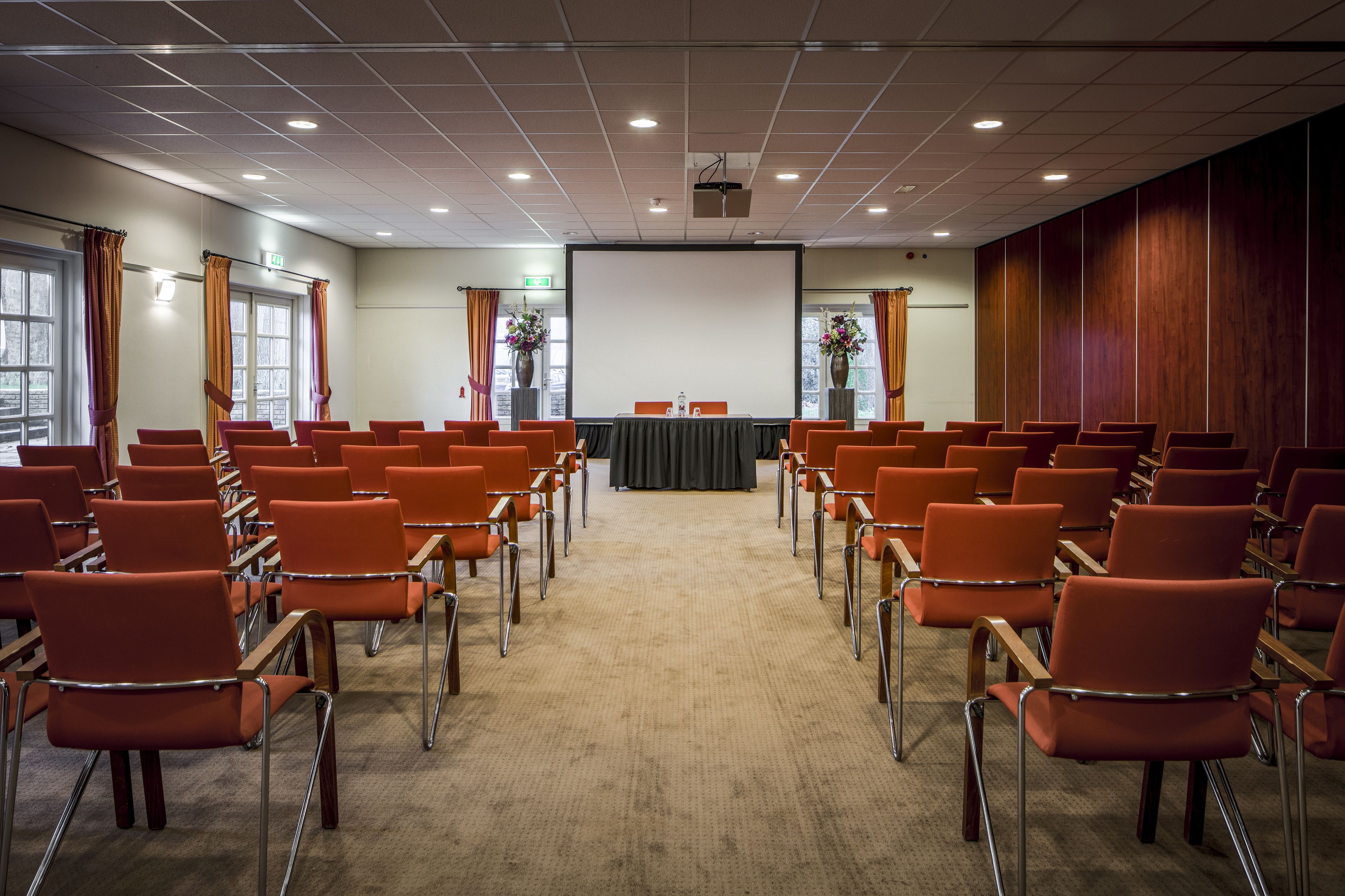 Een vergaderzaal met rijen oranje stoelen, een projectiescherm en een tafel met een zwarte tafelkleed.