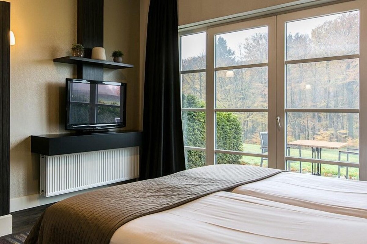 Ein Schlafzimmer mit einem großen Bett, einem Fernseher auf einem schwarzen Regal, einem Fenster mit Blick auf Bäume und einem Heizkörper unter dem Regal.