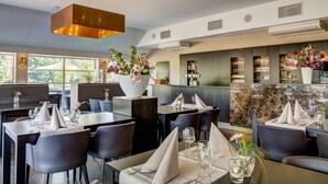 A restaurant with dark-toned tables and chairs, a large white vase with flowers, and a bar area with shelves of glasses and bottles.