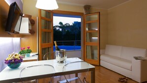 Una habitación con una mesa blanca, sillas, un sofá blanco, un televisor montado en la pared y una ventana con vistas a palmeras.