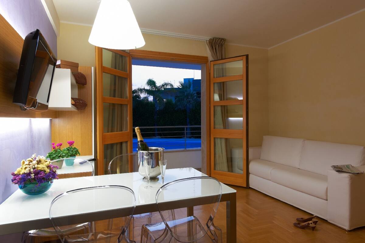A room with a white table, chairs, a white sofa, a TV mounted on the wall, and a window with a view of palm trees.