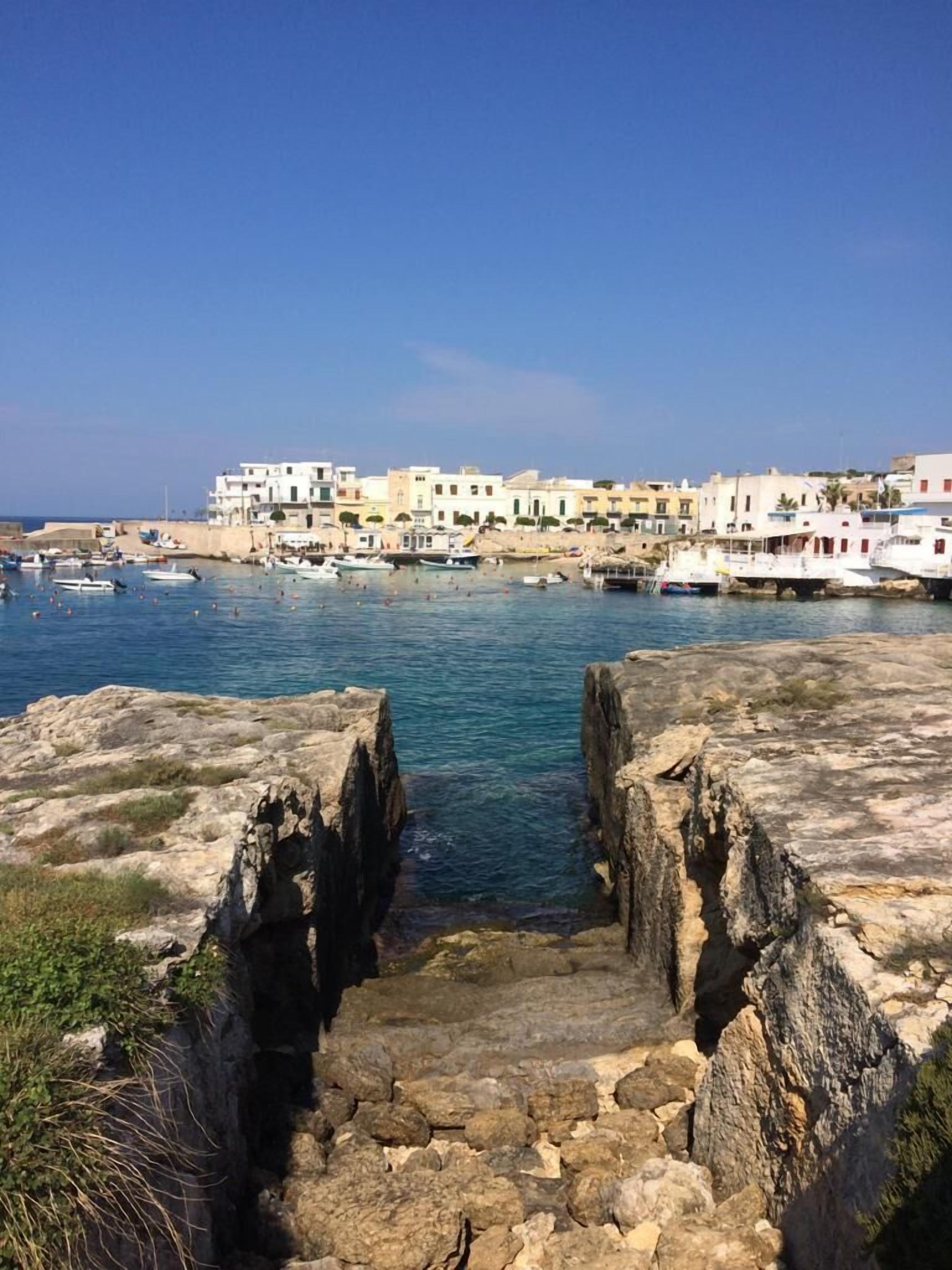 A coastal town with stone steps leading to a harbor, surrounded by buildings with balconies and a clear blue sky.