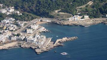Vista aérea de un pueblo costero con edificios a lo largo de la costa, un rompeolas rocoso y un castillo en el fondo.