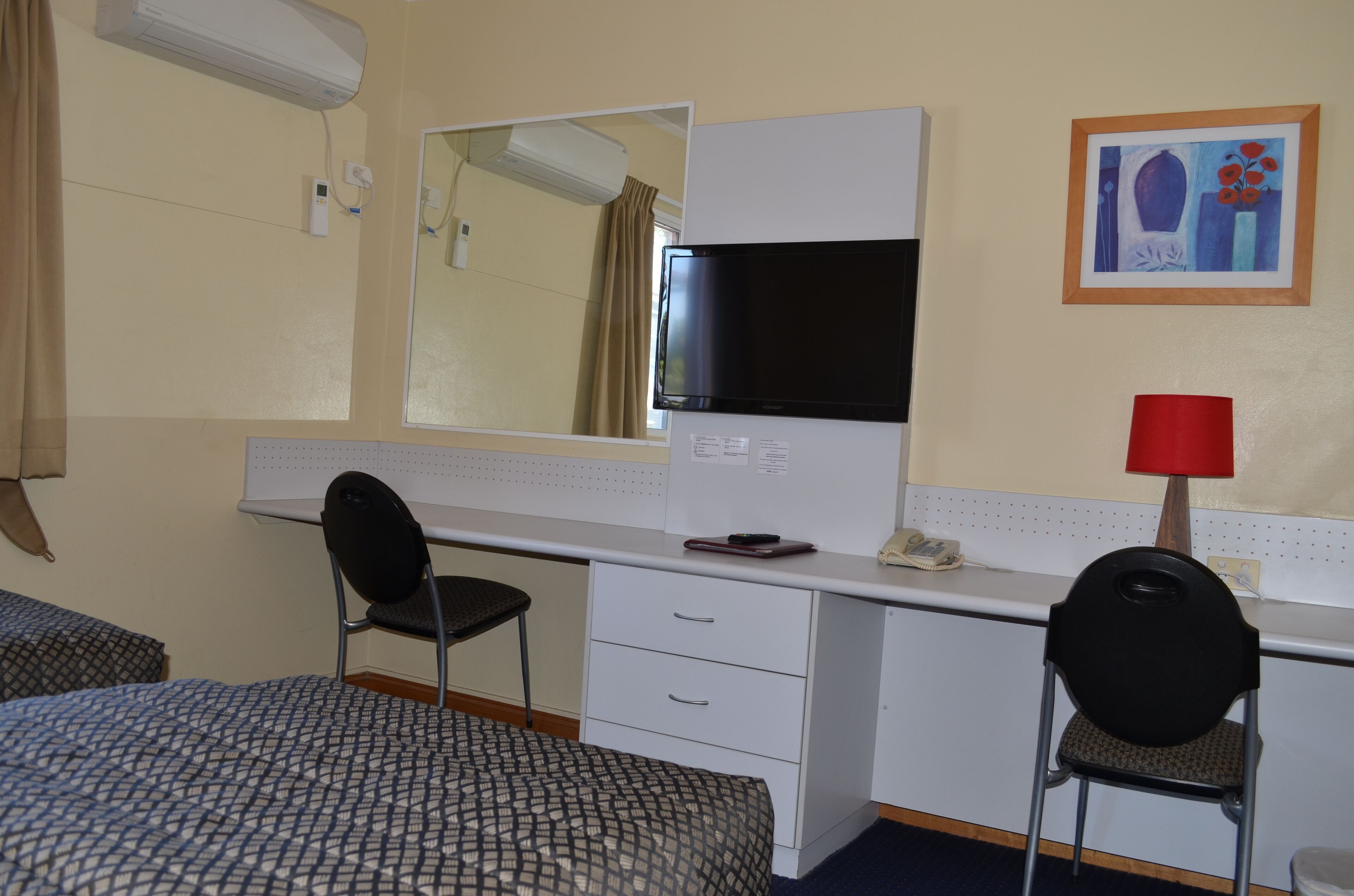Une chambre d’hôtel avec un lit simple, un bureau avec une chaise, un miroir, un téléviseur à écran plat, une lampe rouge et un tableau au mur.