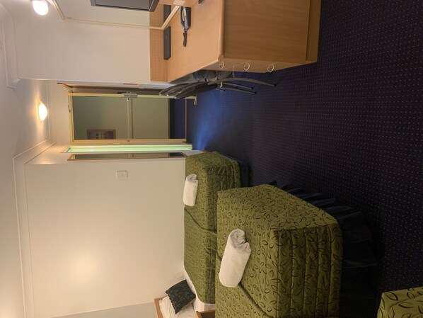 Habitación de hotel compacta con dos camas individuales, un escritorio pequeño con lámpara, un espejo y una alfombra azul.