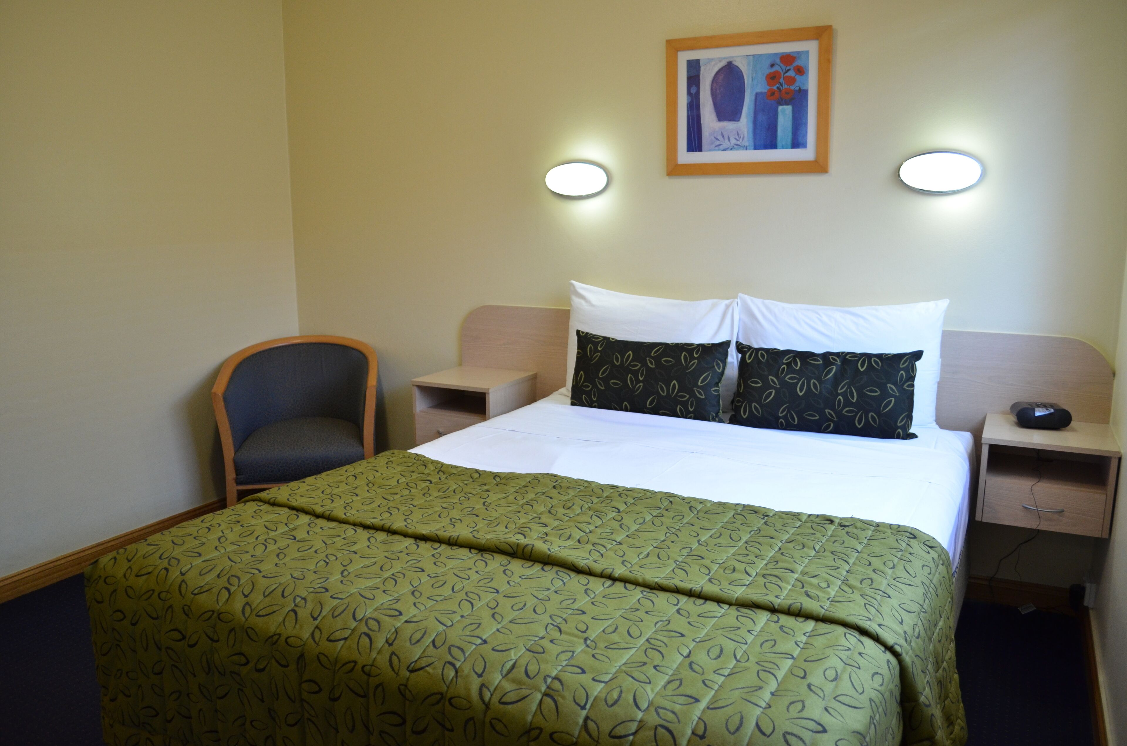 A hotel room with a bed, a chair, a nightstand, and a wall-mounted picture.