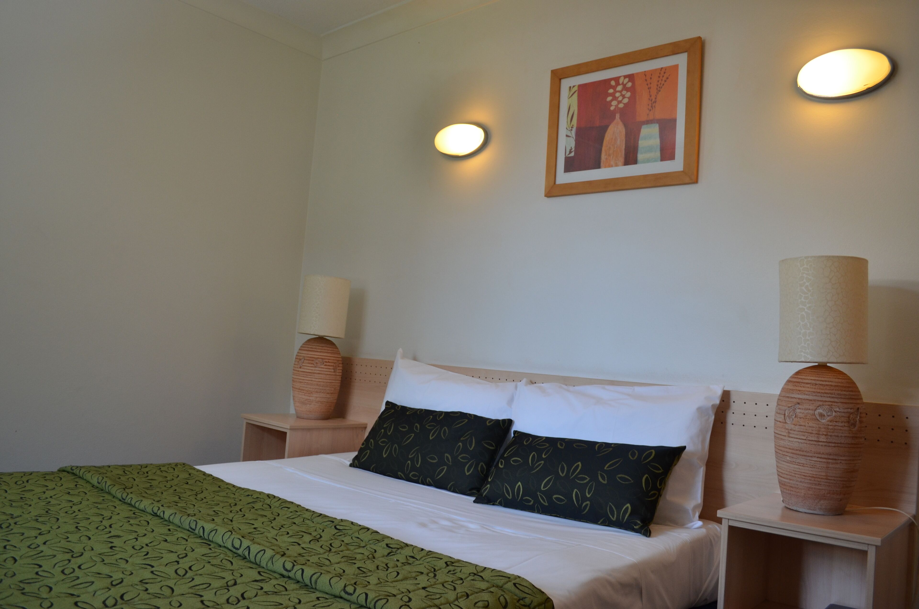 A neatly made bed with a green patterned bedspread, two pillows, and a headboard. There are two bedside tables with lamps and a framed picture on the wall above the bed.