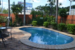 A swimming pool with a curved edge, surrounded by a brick patio, palm trees, and a fence. - Airport Motel (Hamilton)