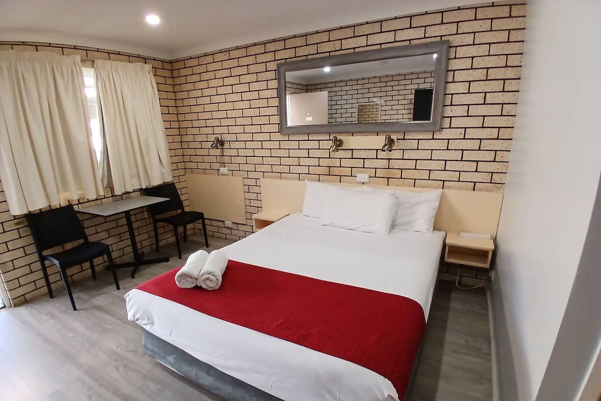 Habitación de hotel con pared de ladrillo, cama con ropa de cama blanca y roja, un escritorio con silla y una mesita de noche con lámpara.