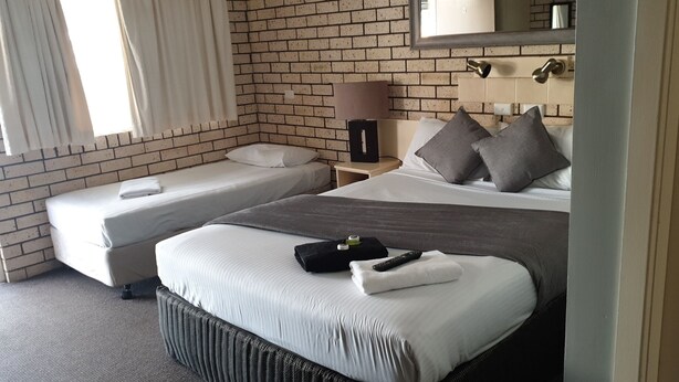 Habitación de hotel con pared de ladrillo, una cama individual con ropa de cama gris, una cama más pequeña, una mesita de noche con lámpara y una ventana con cortinas.