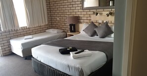 A hotel room with a brick-patterned wall, a single bed with grey bedding, a smaller bed, a nightstand with a lamp, and a window with curtains.