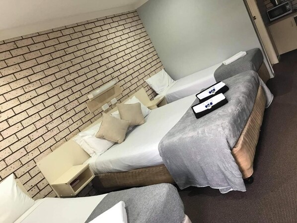 A hotel room with a brick-patterned wall, a bed with a grey blanket, two pillows, a bedside table with a lamp, and a small bedside table with a book.