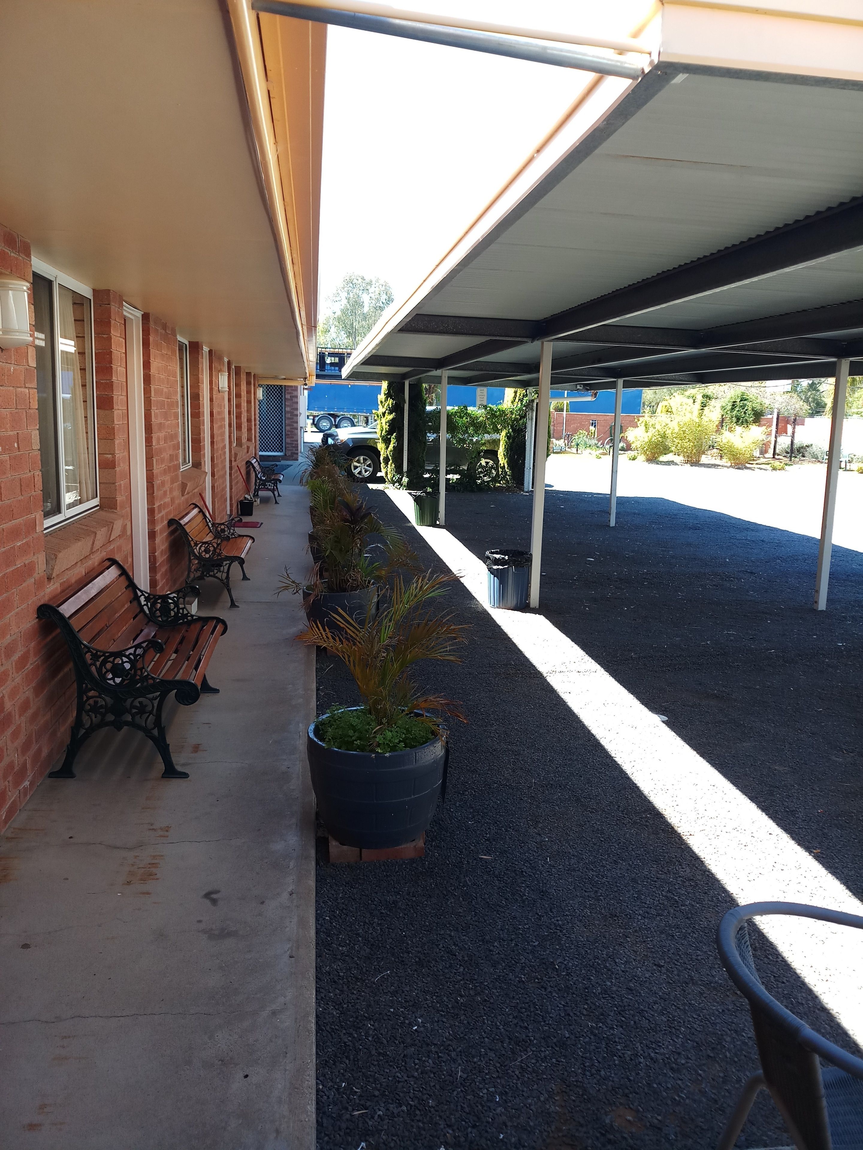 A covered outdoor area with benches and potted plants, a brick building, and a blue structure in the background.