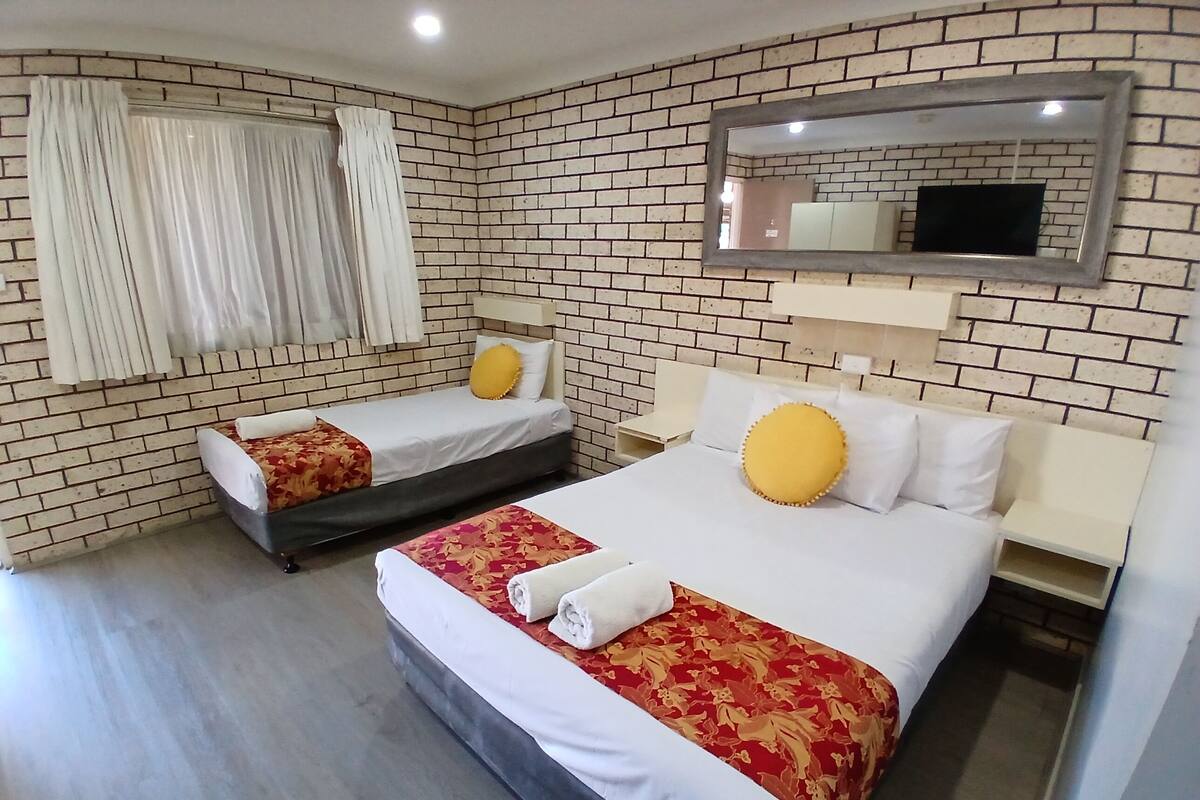 Habitación de hotel con dos camas, pared de ladrillo, espejo y ventana con cortinas.