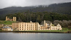A historic stone building with multiple wings, situated by a body of water, with a forested hill in the background. - Alexanders' Of Richmond (Richmond)