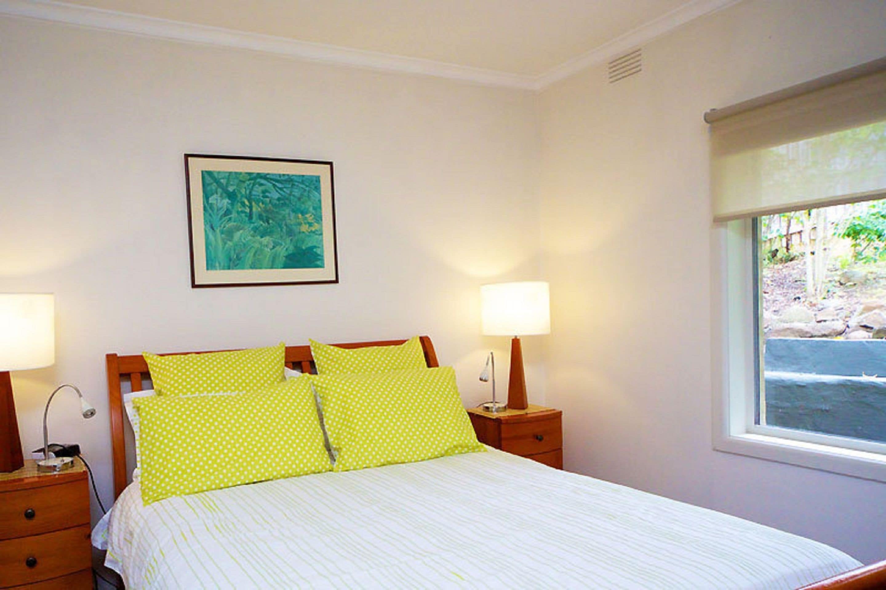 Una cama bien hecha con dos almohadas, una mesita de noche con una lámpara, y una ventana con vistas al exterior.