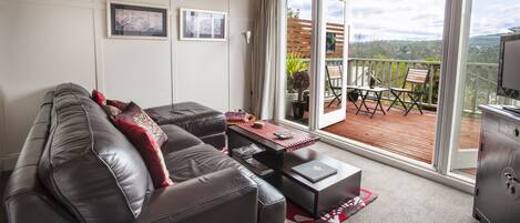 Un salon moderne avec un canapé en cuir, une table basse et une porte-fenêtre coulissante donnant sur un balcon avec vue sur la campagne.