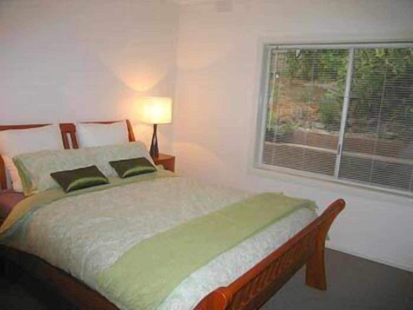 A bedroom with a bed, a nightstand with a lamp, and a window with blinds.