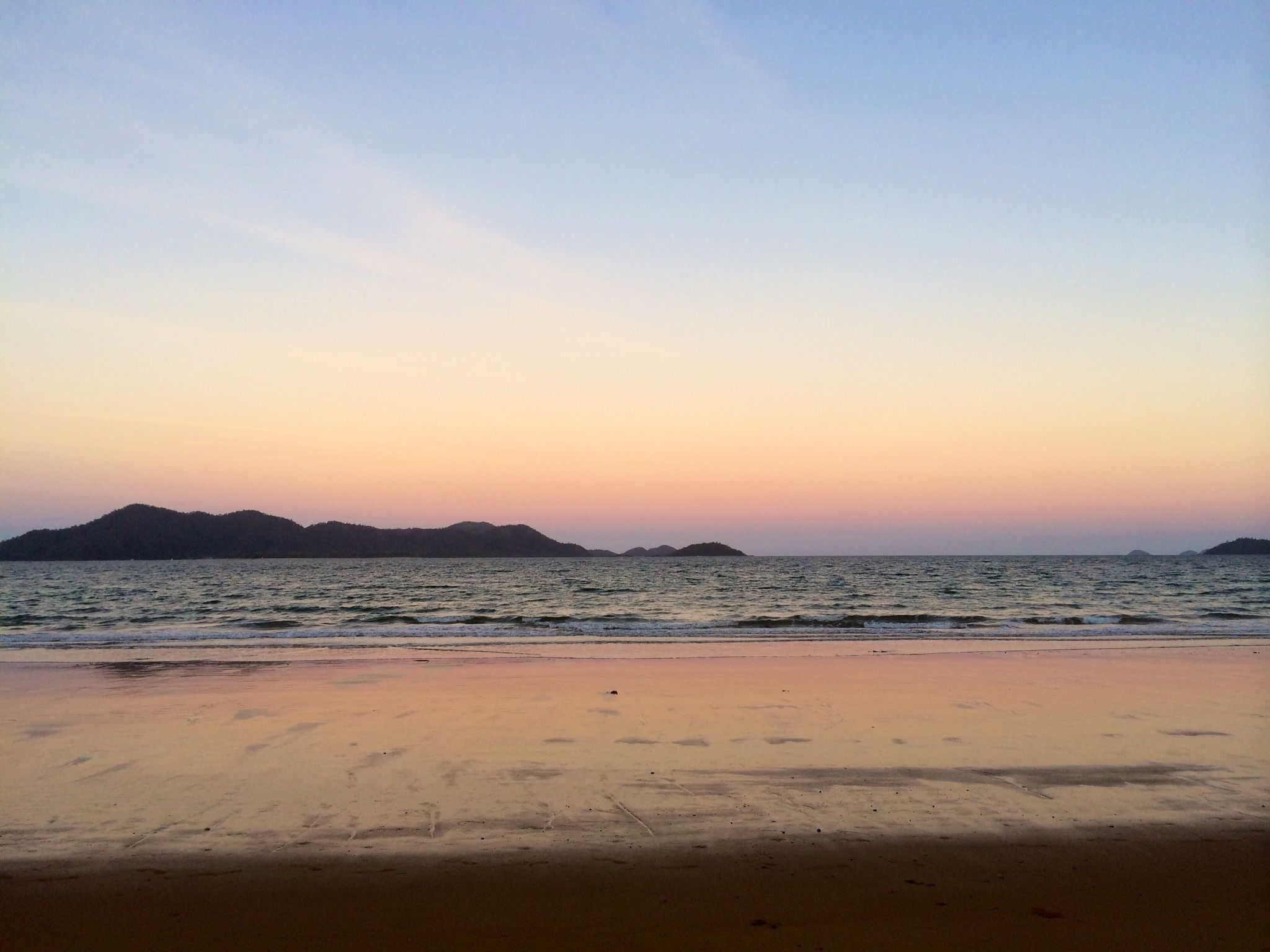 A beach with calm waters and distant mountains during sunset.