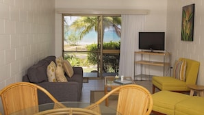 A modern living room with a grey sofa, yellow armchair, and wicker chairs. A flat-screen TV is on a wooden stand, and there's a large window with a view of palm trees and a beach. - Apollo Jewel Beachfront Apartments (South Mission Beach)