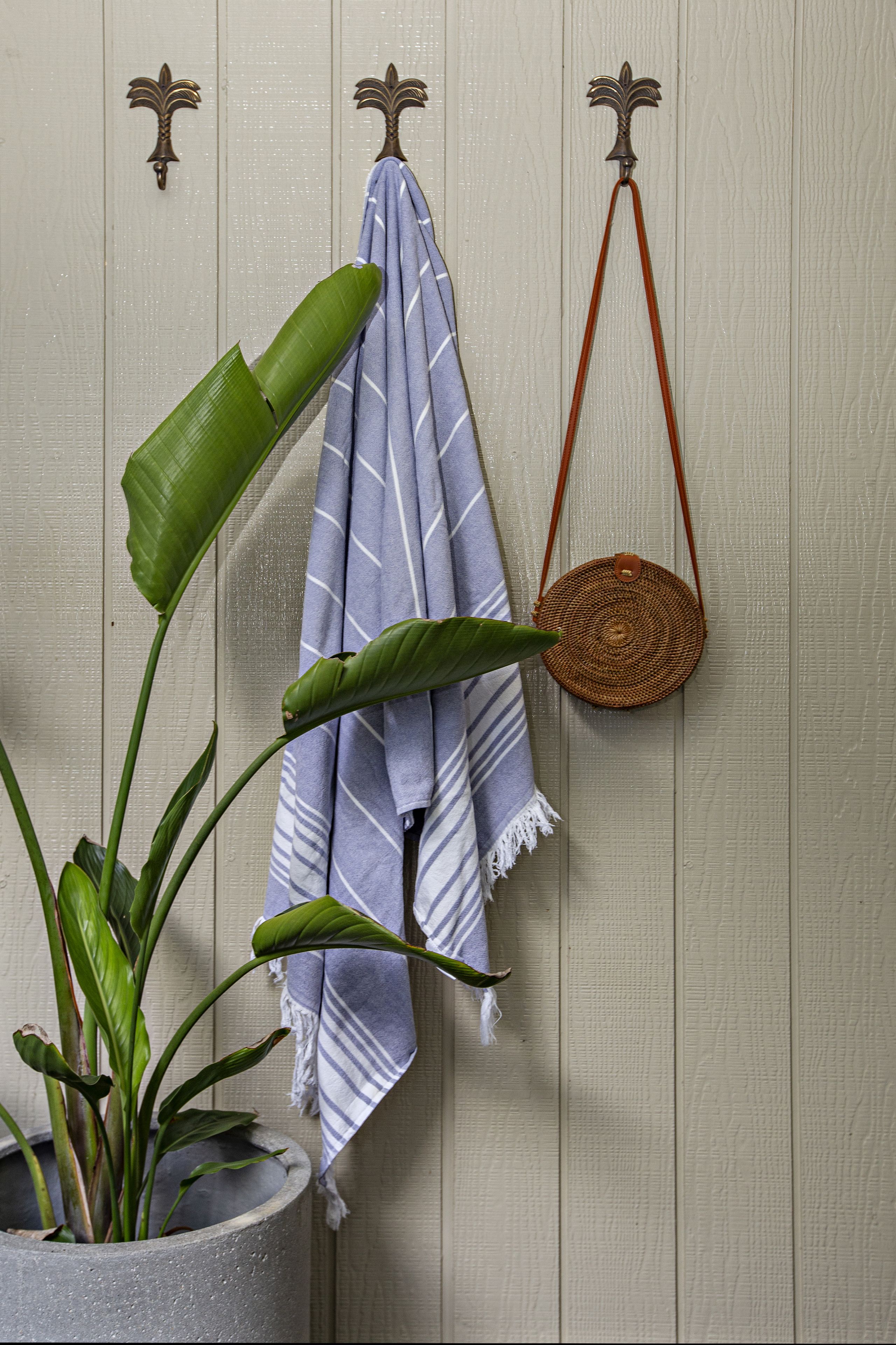 Une plante en pot aux grandes feuilles, une serviette à rayures et un panier tressé suspendu à un mur grâce à des crochets décoratifs.