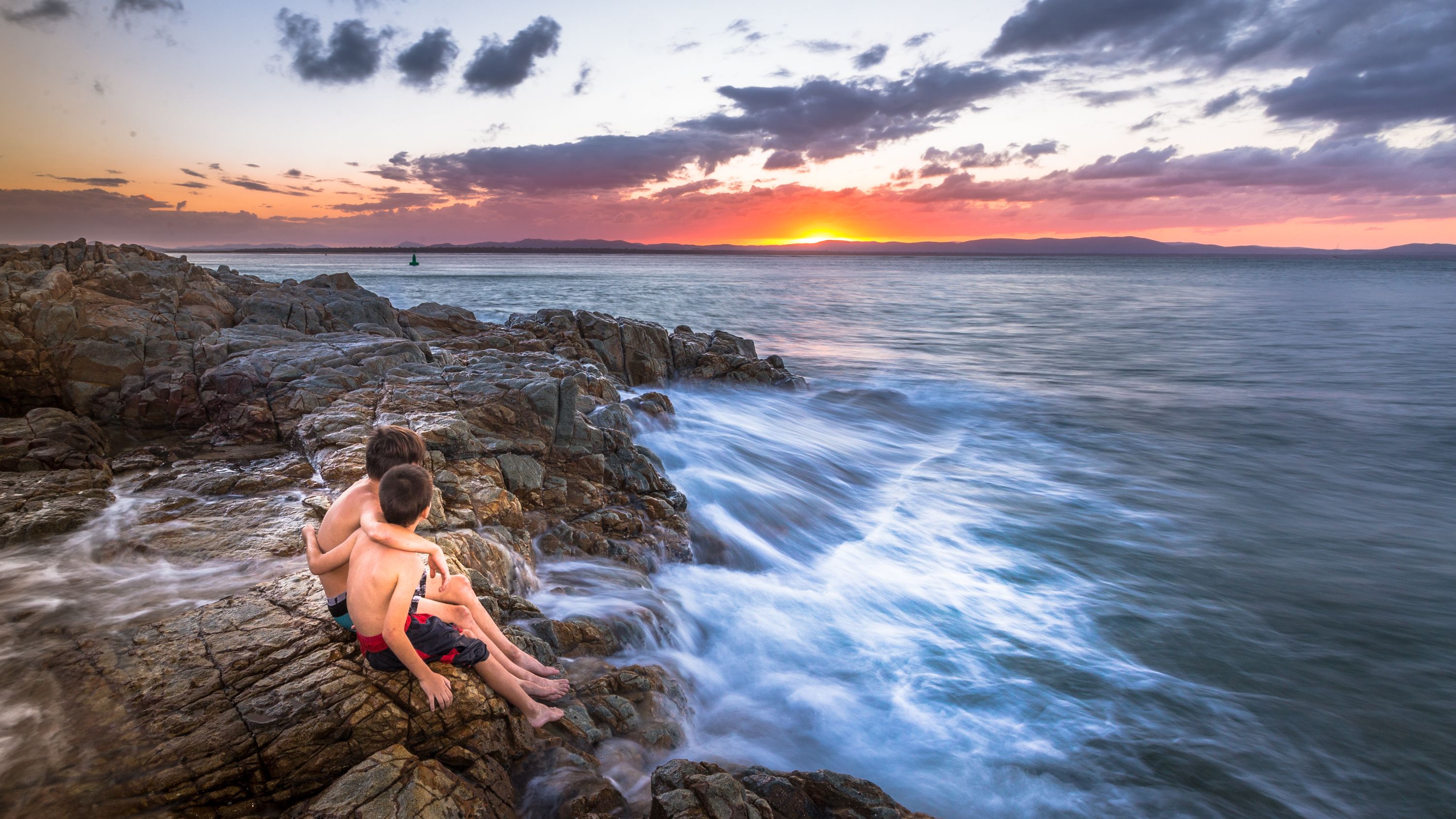 A sunset view over rocky coastline with two individuals sitting on rocks.