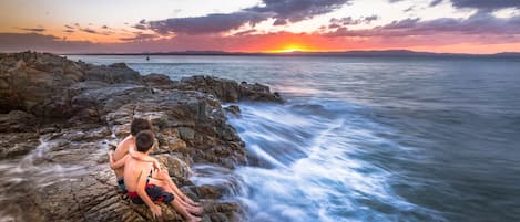 A sunset view over rocky coastline with two individuals sitting on rocks.