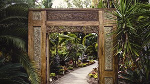 Un arco de madera ornamentado con tallas intrincadas, que conduce a un exuberante sendero de jardín rodeado de diversas plantas y flores tropicales.