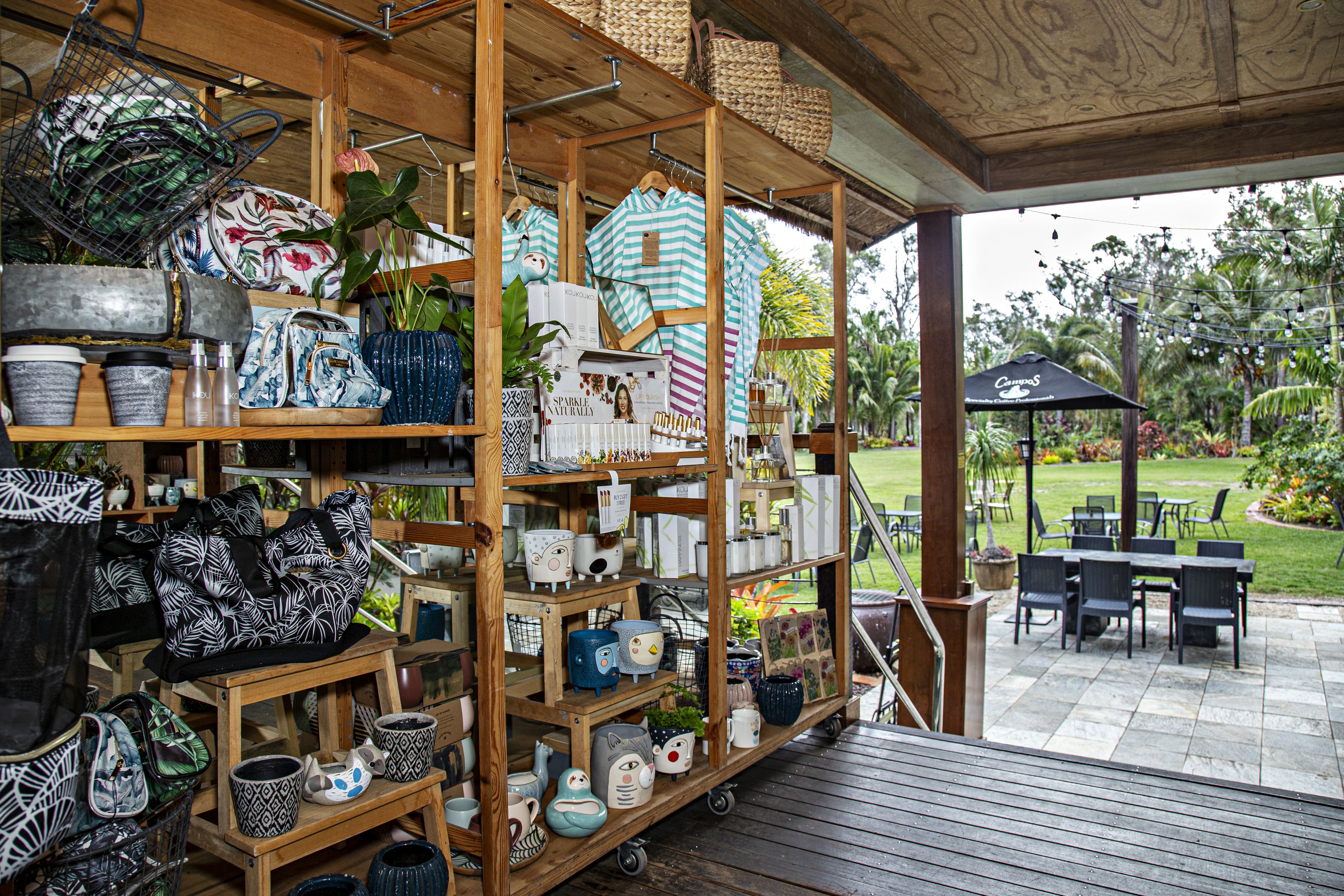 Een overdekt buitengebied met houten planken waarop diverse keramieken objecten staan, en een terras met tafels en stoelen.