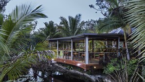 Un espacio de estar al aire libre con techo y piso de madera sobre una masa de agua, rodeado de vegetación tropical.