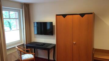 Une pièce avec un téléviseur fixé au mur, une armoire en bois, un bureau avec une chaise et une fenêtre avec des rideaux.