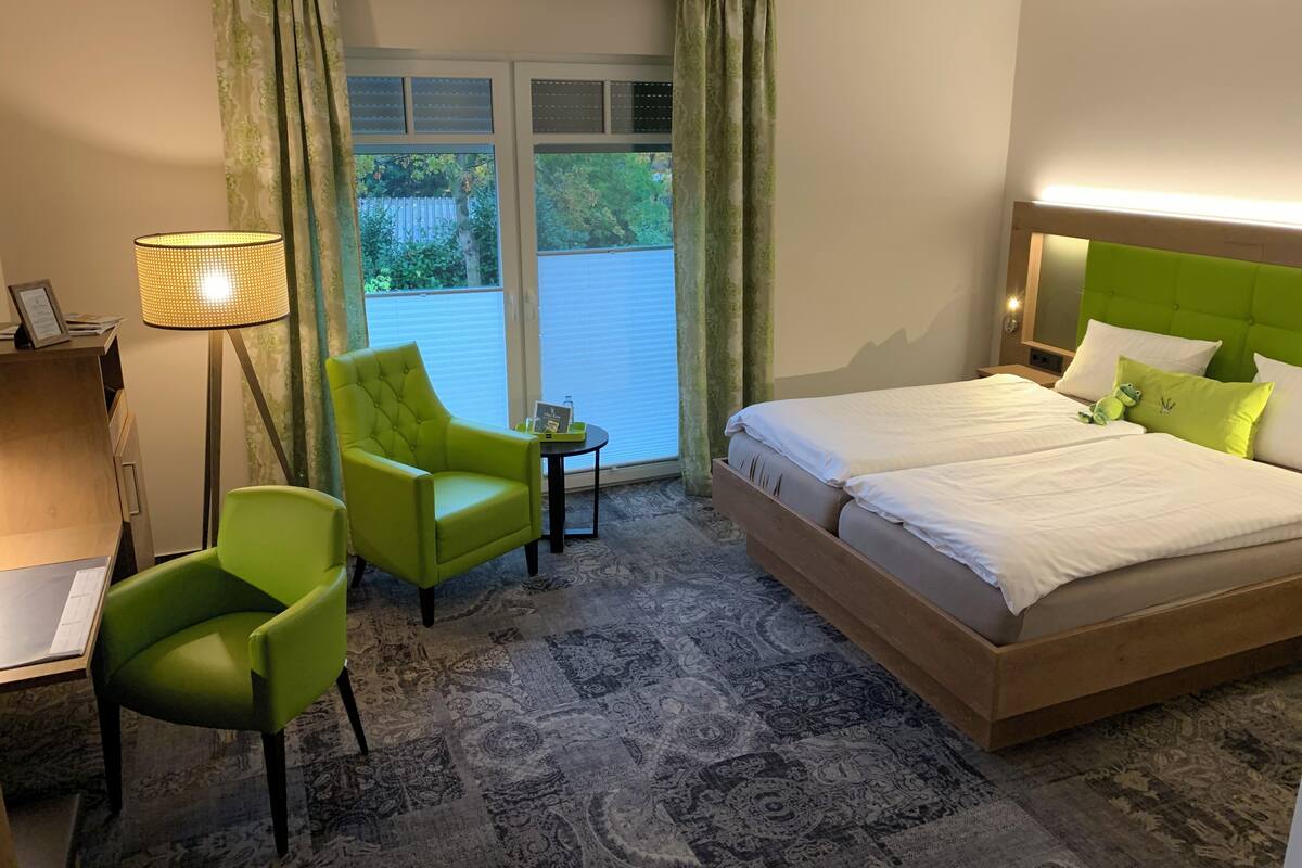 A hotel room with a bed, two green chairs, a desk, a lamp, and a view of the outdoors.