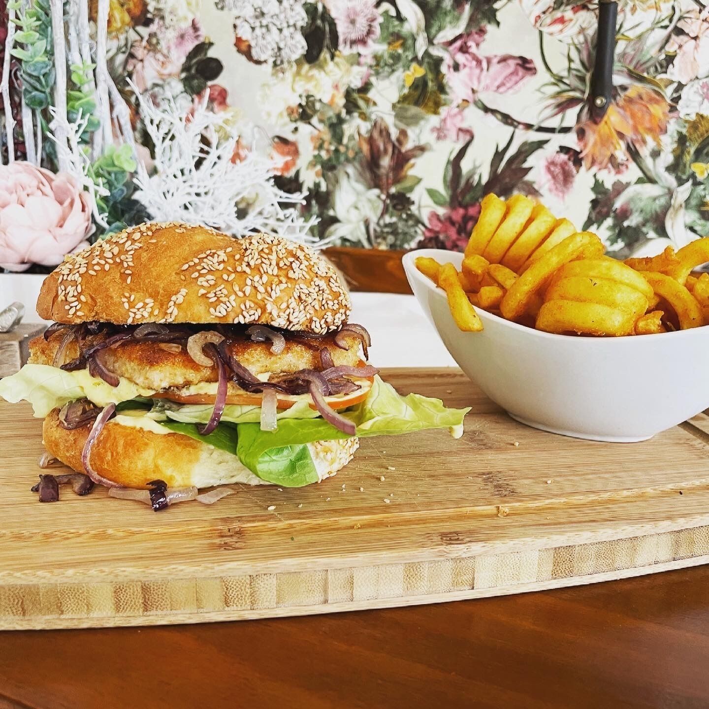 Un sándwich con pan de sésamo, lechuga y cebolla, junto a un bol de papas fritas sobre una tabla de madera.