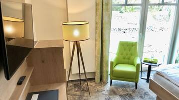 A hotel room with a bed, a green armchair, a floor lamp, a desk with a remote control, and a window with a view of the outdoors.