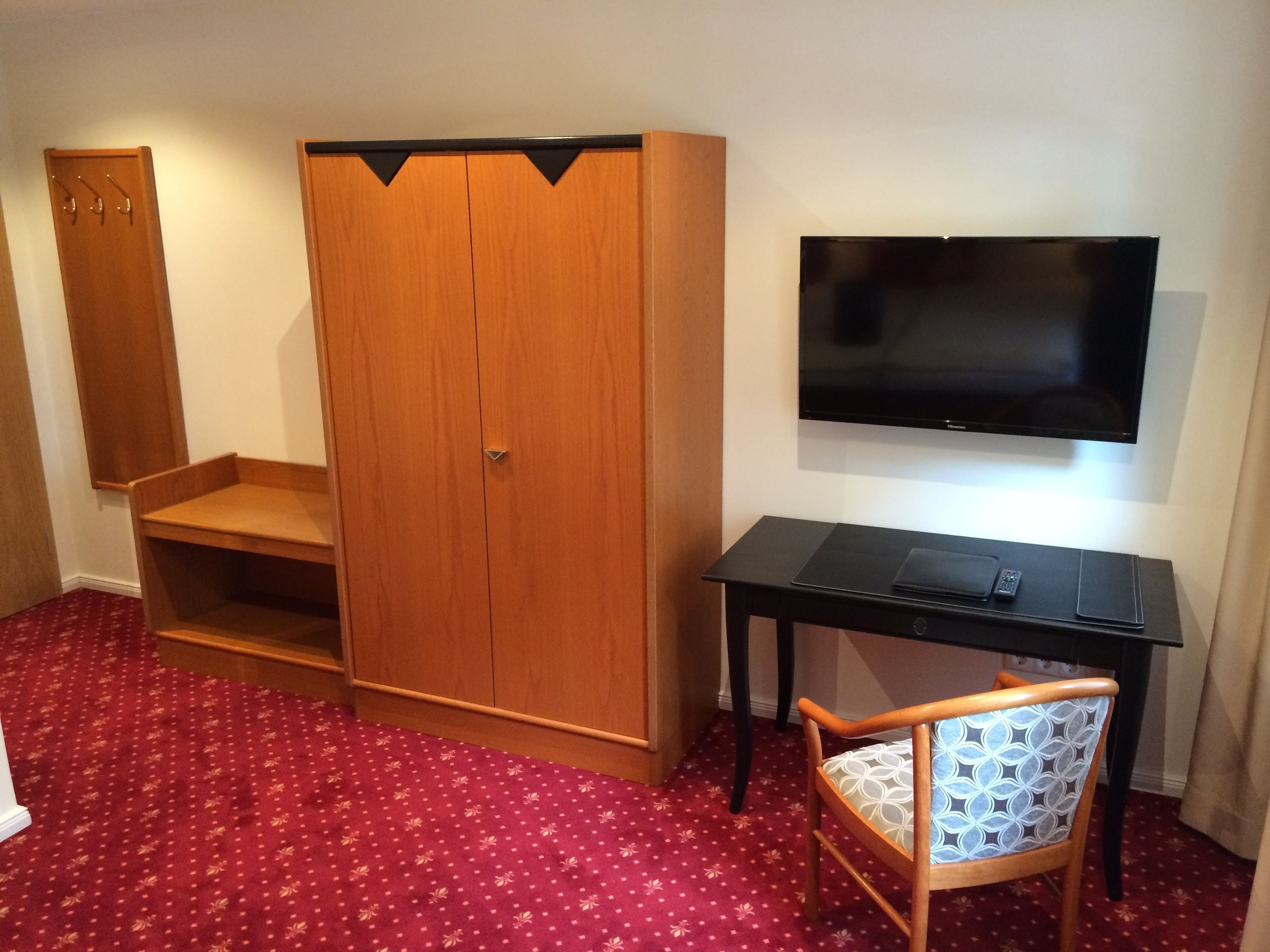 Ein Zimmer mit einem Holzschrank, einem an der Wand montierten Fernseher, einem Schreibtisch mit Stuhl und einem roten Teppich mit Muster.