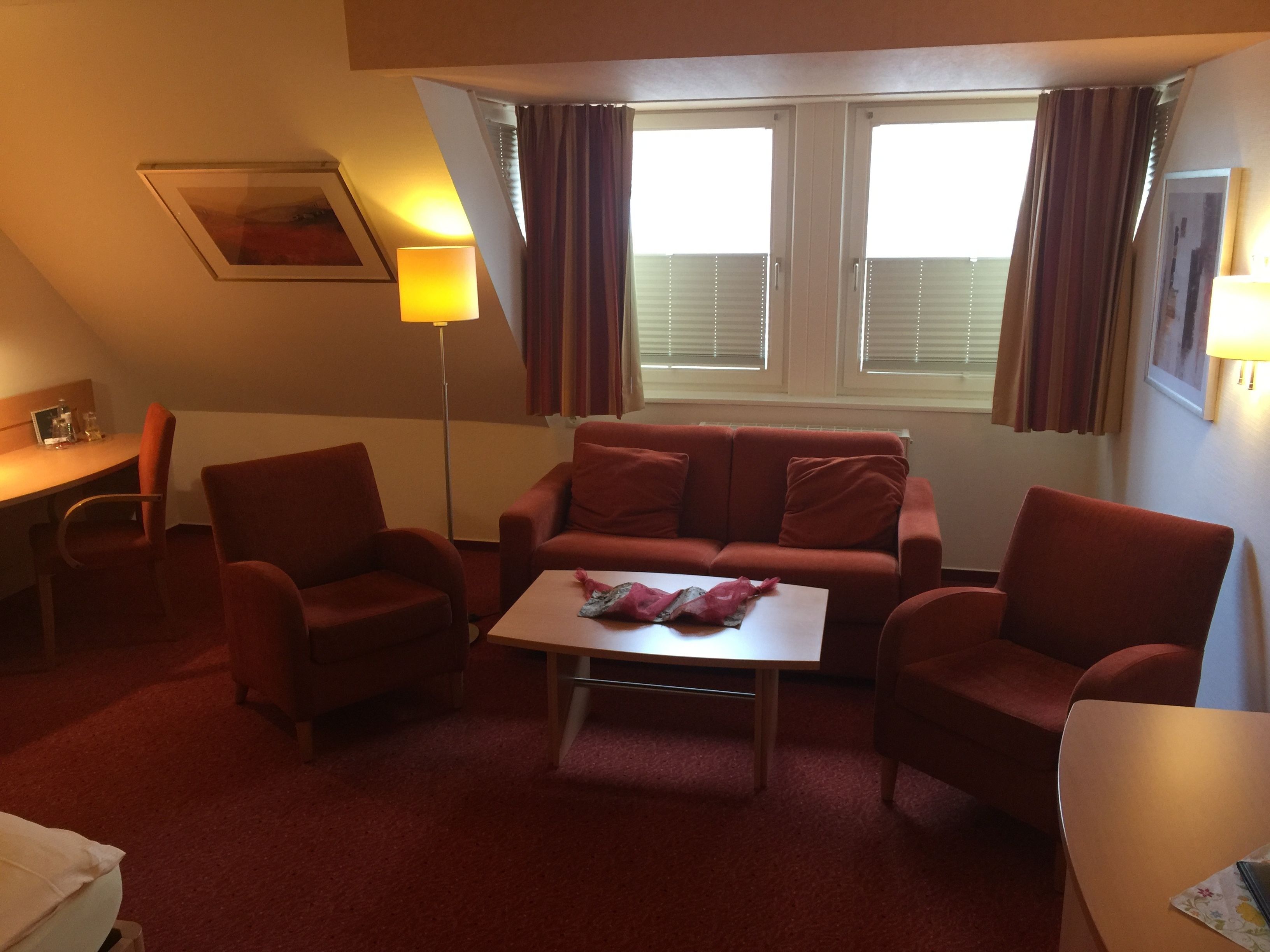 Un salon chaleureux avec un canapé rouge, une table basse et une lampe. Il y a une fenêtre avec des rideaux et un tableau encadré accroché au mur.