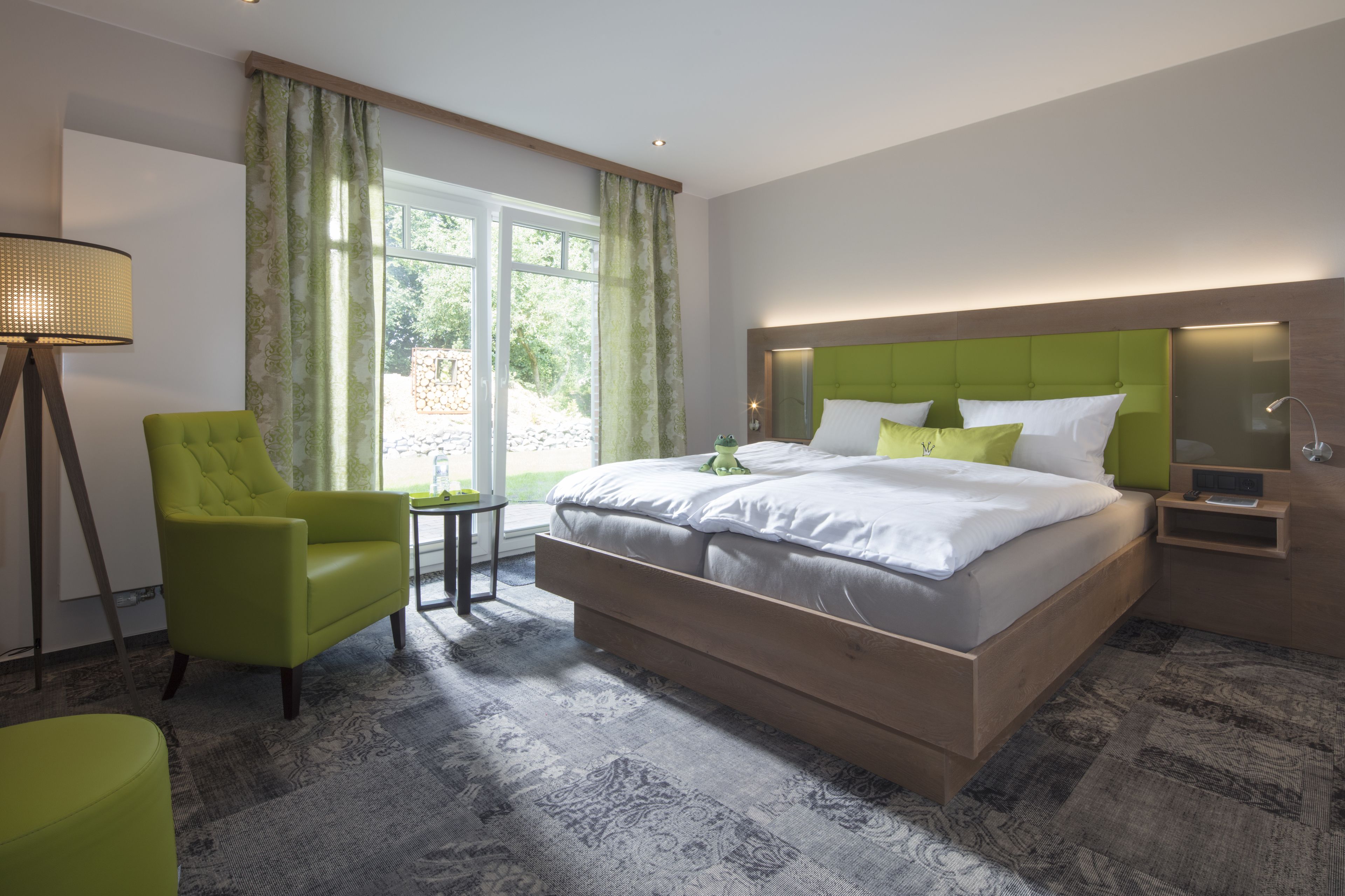 Habitación de hotel moderna con una cama grande, un sillón verde, una mesita pequeña y vistas al exterior con zonas verdes.