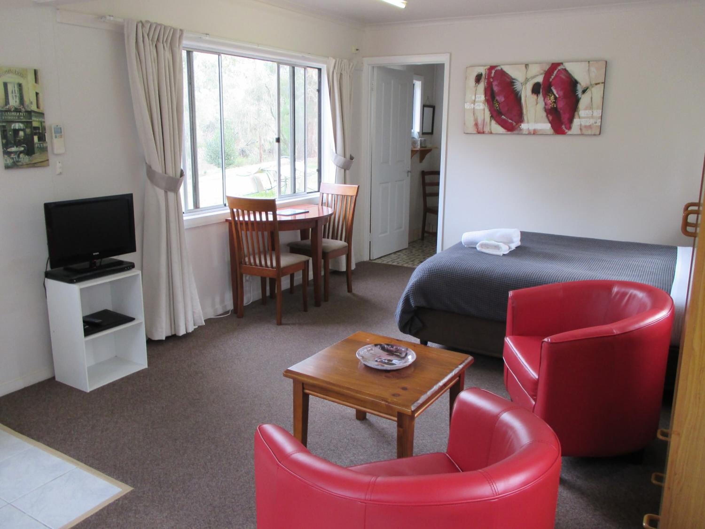 A room with a bed, a TV, a dining table with chairs, a red chair, and a painting on the wall.