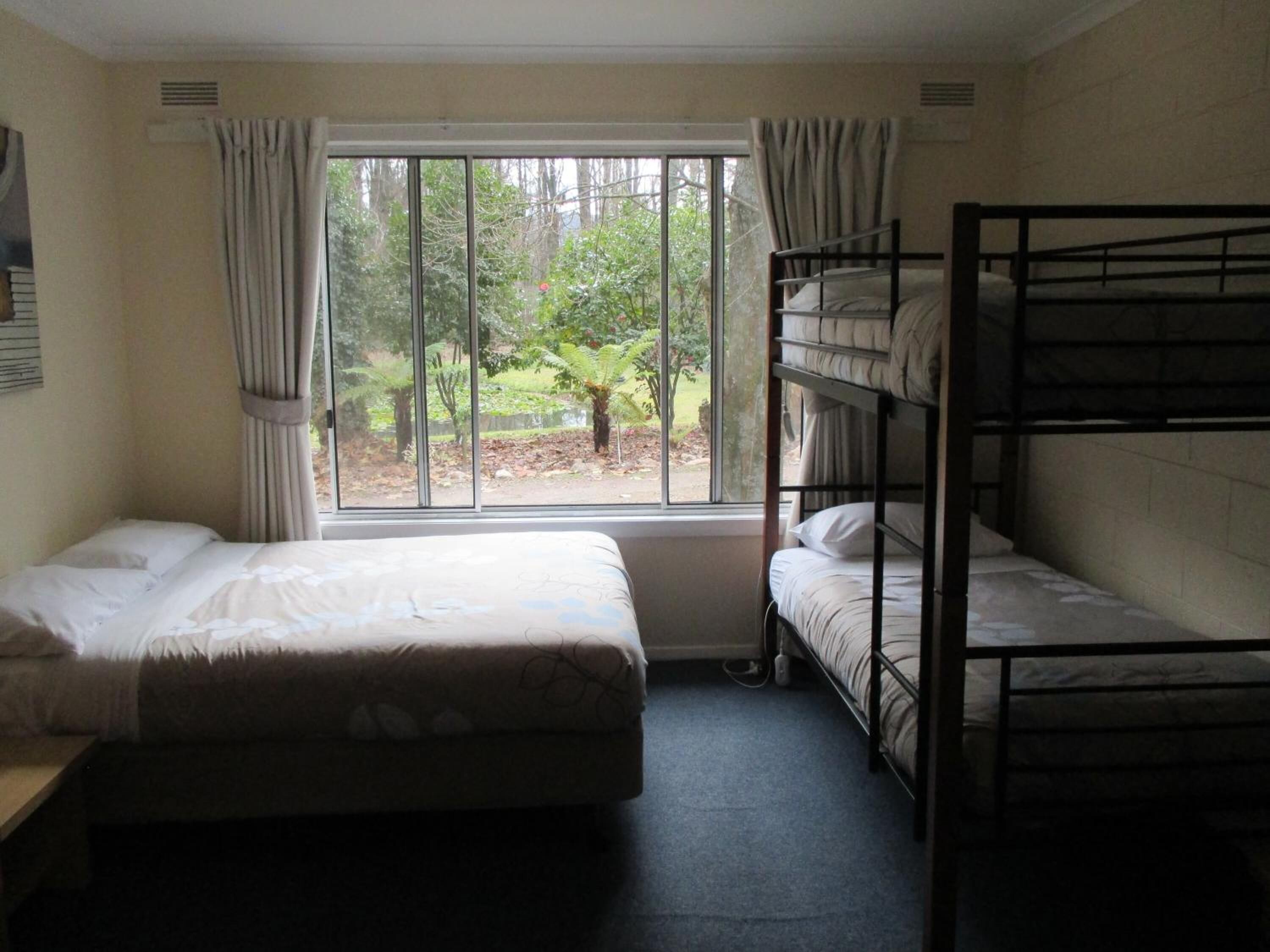 A dormitory room with two single beds and a bunk bed, large windows with curtains, and a view of trees outside.