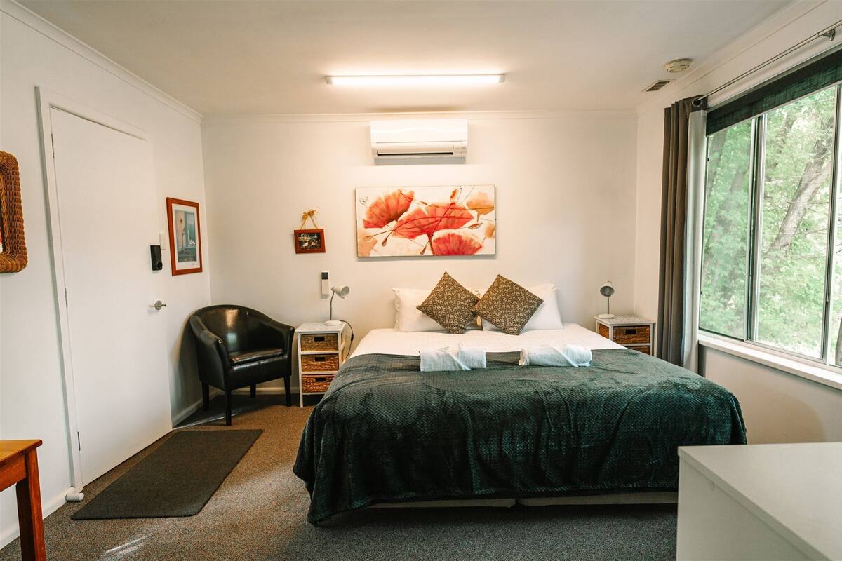 A bedroom with a bed, a chair, a bedside table, an air conditioner, and a window with a view of trees.