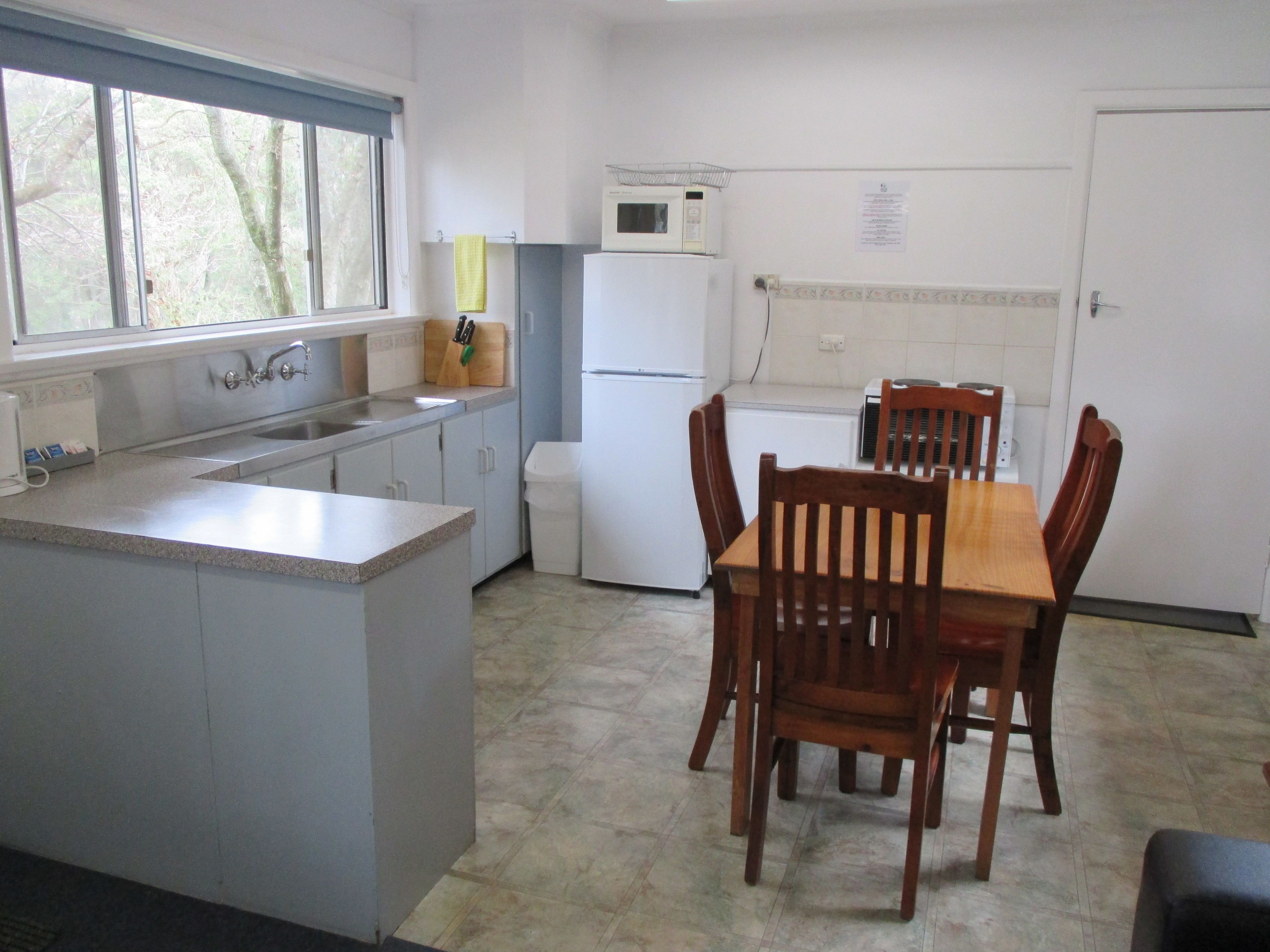 A compact kitchen with a central island, a dining table with chairs, a refrigerator, a microwave, and a window with a view of trees.