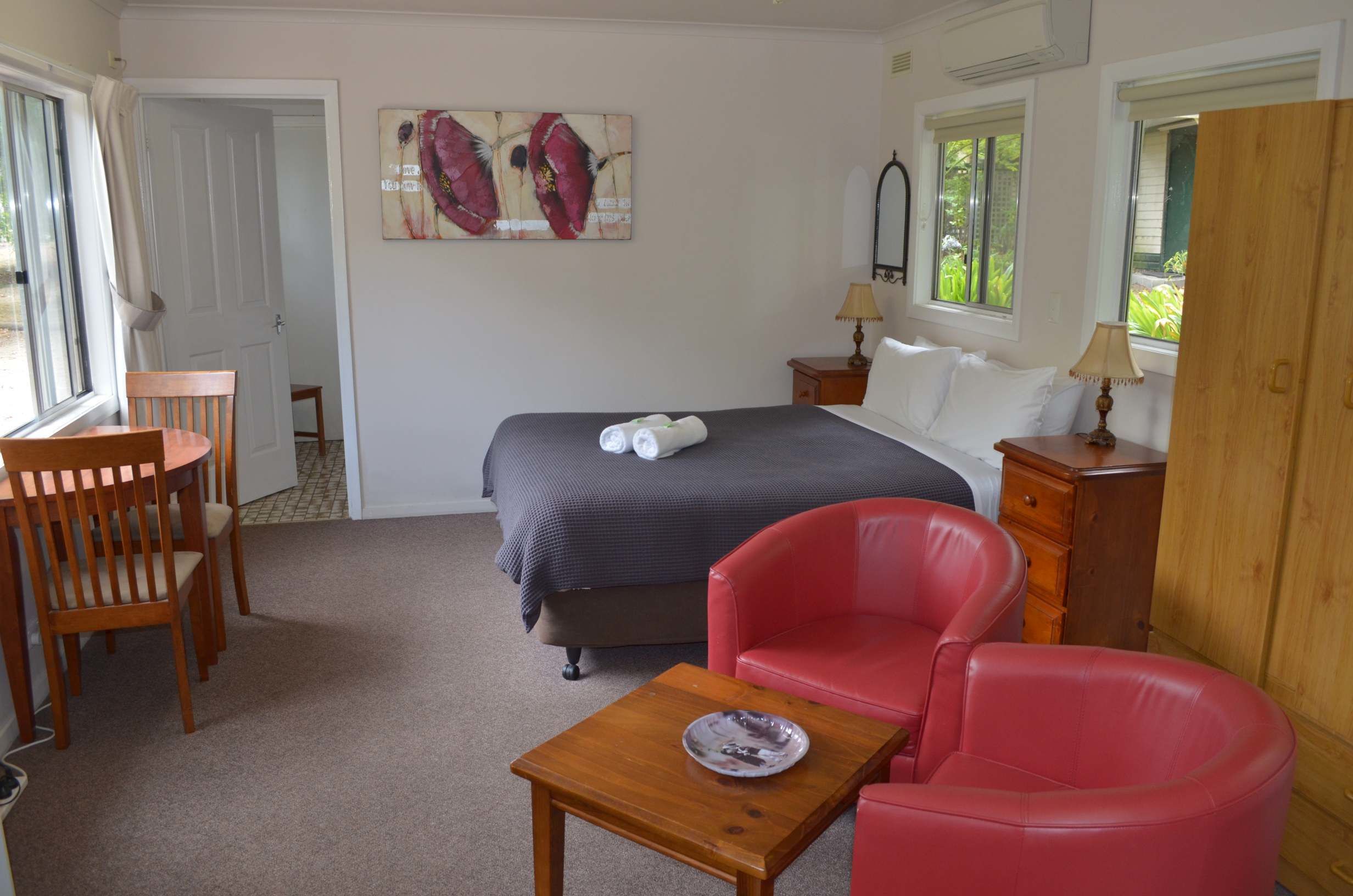 Een slaapkamer met een bed, een rode stoel, een houten tafel, een schilderij aan de muur en een raam met zonwering.