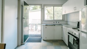 Ett kompakt kök med vita skåp, en spis och ett kylskåp. Det finns ett fönster som släpper in naturligt ljus och en dörr som leder ut till ett utomhusområde med ett bord och stolar.