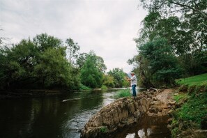 A person fishing by a river with dense greenery and trees in the background.