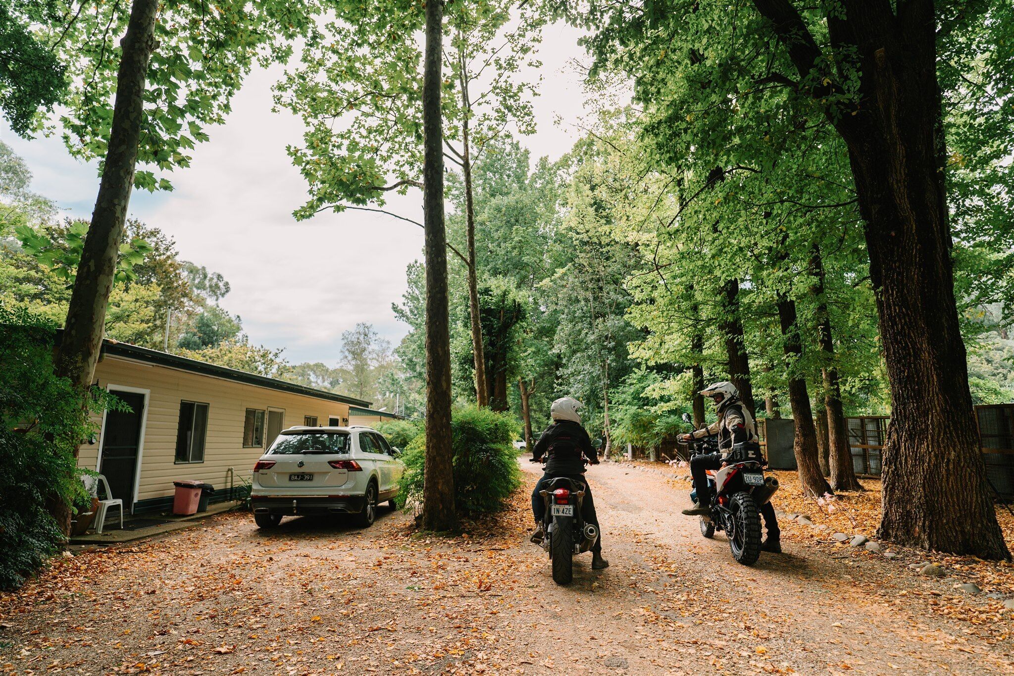 Un chemin de terre avec deux motos garées sur le bord, une voiture blanche et une caravane dans une zone boisée.