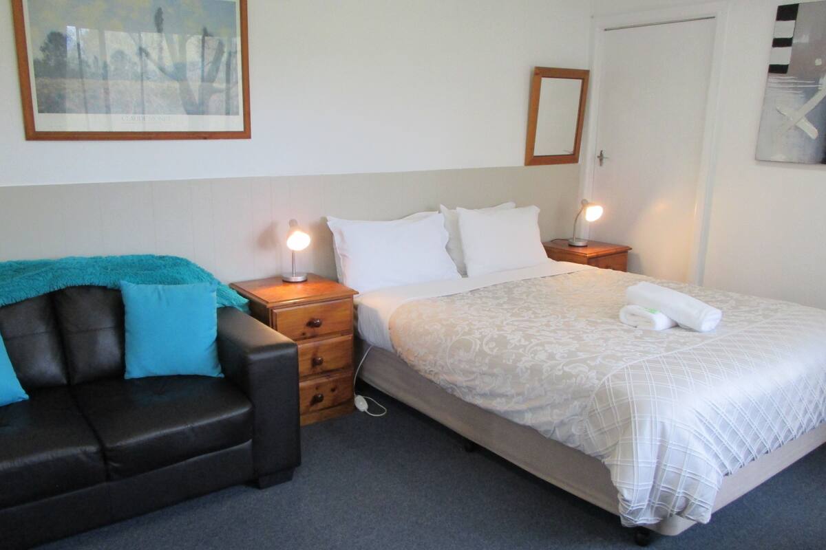 A neatly arranged bedroom with a bed, a nightstand, a lamp, a couch, and a painting on the wall.