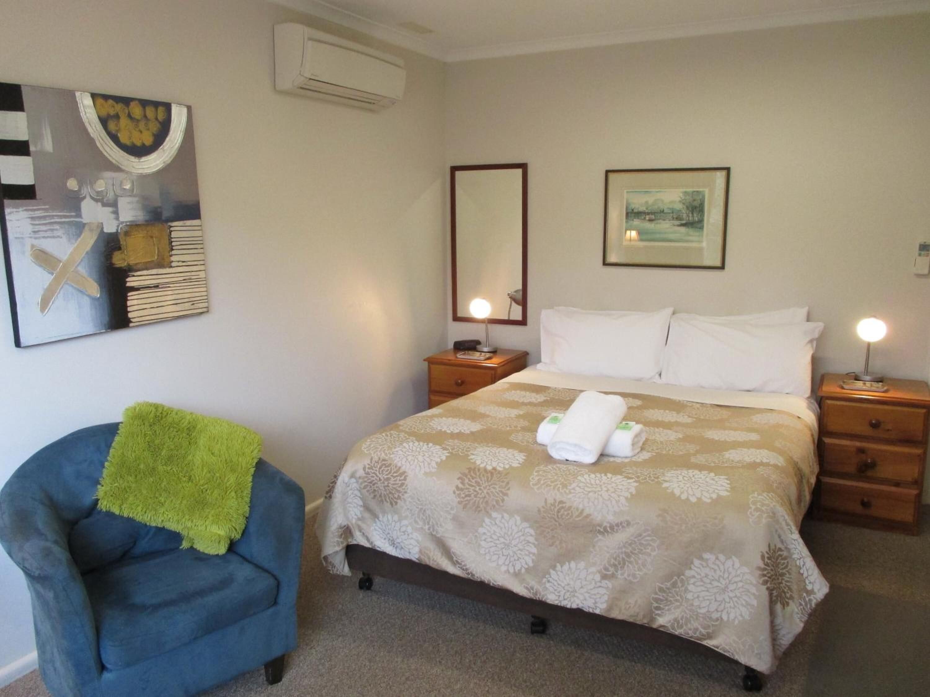 Una habitación de hotel con una cama, dos mesitas de noche con lámparas, una silla con un plaid verde y obras de arte en las paredes.