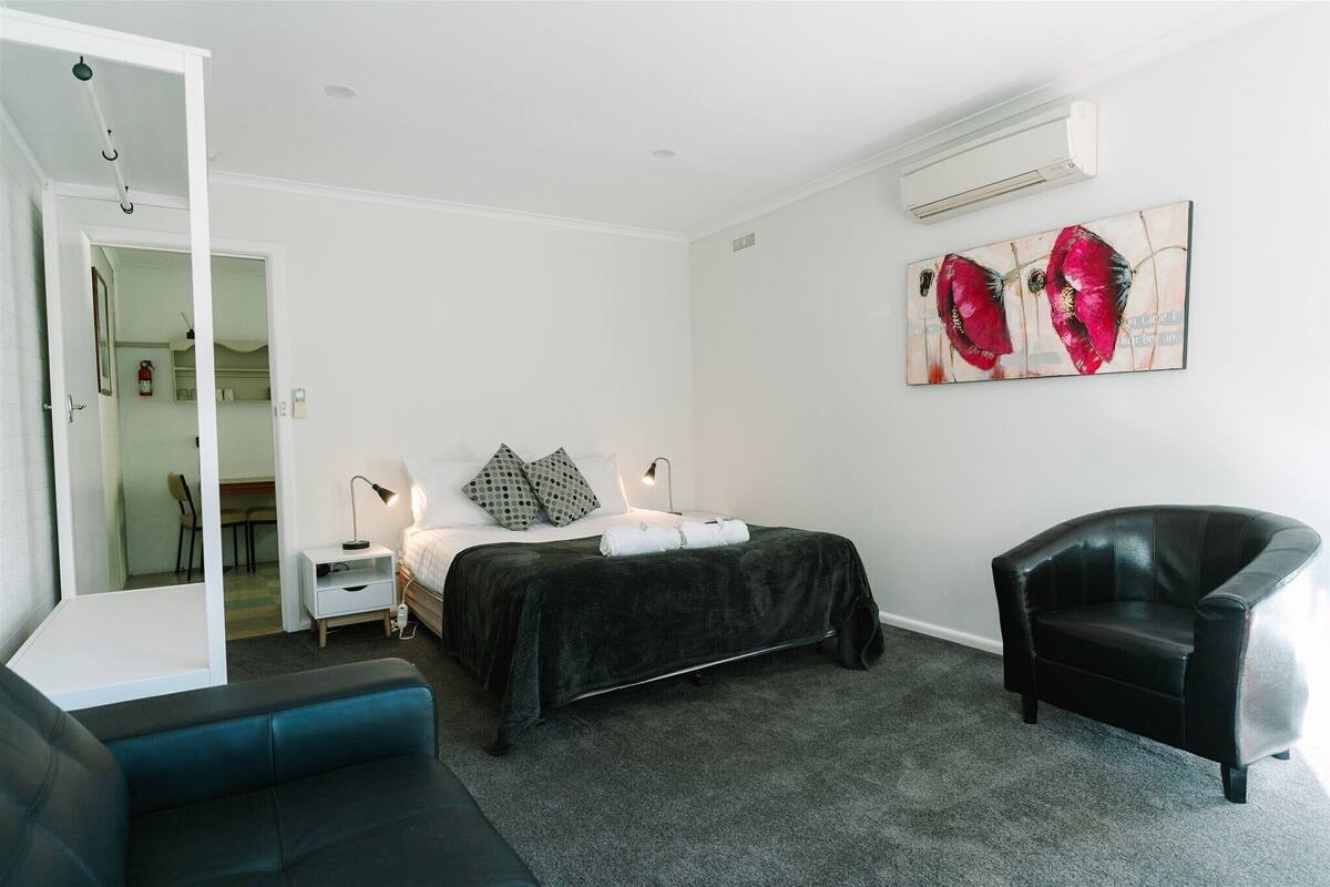 A bedroom with a bed, a chair, a bedside table, and a wall-mounted air conditioner.